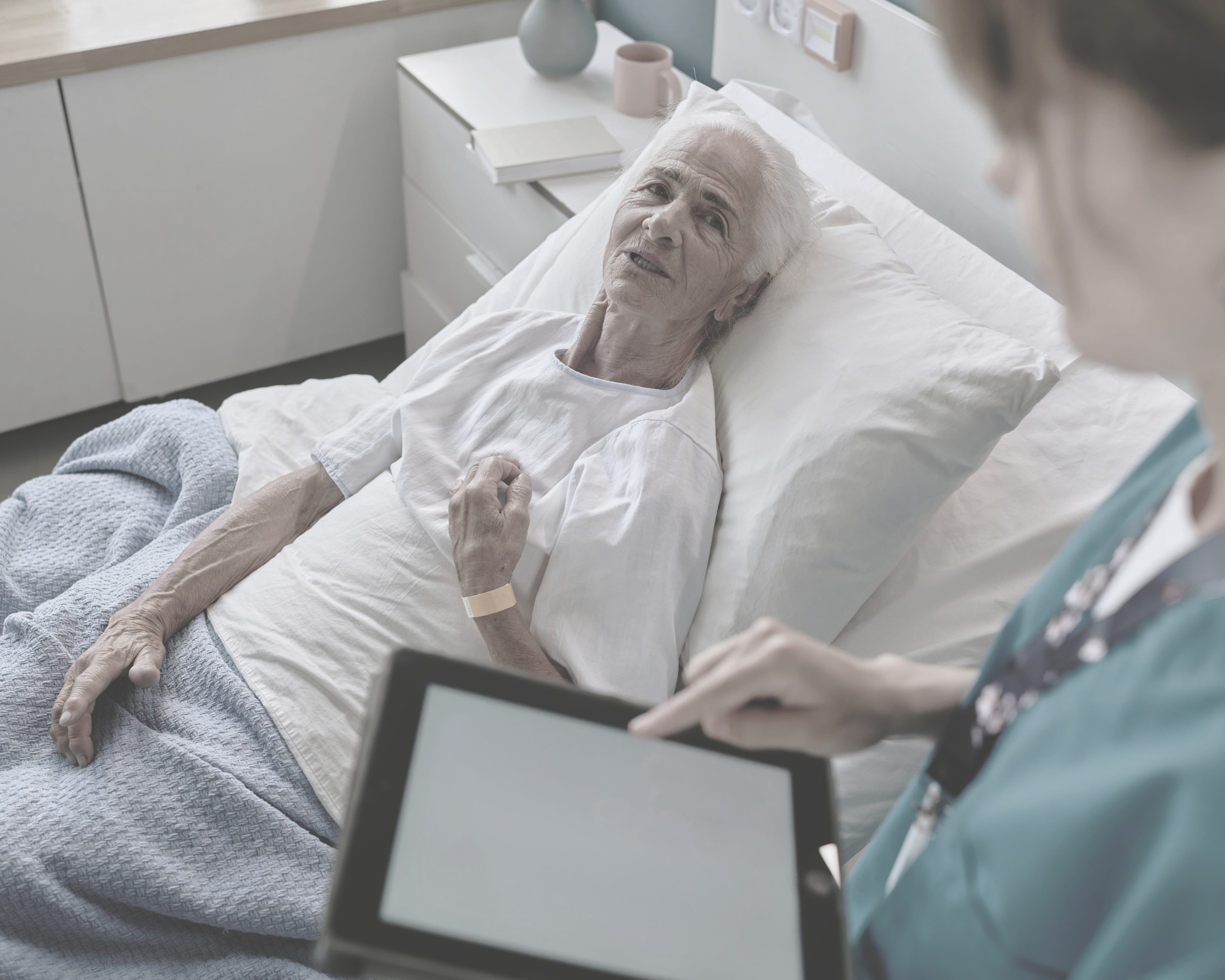 A nurse reviews care details on a tablet at the bedside of a recovering elderly patient.